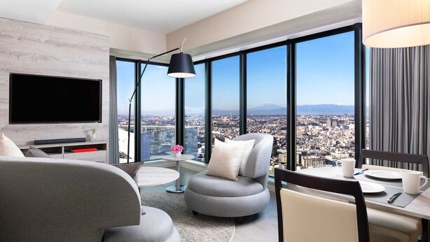 A modern hotel room with a large window offering a city view, a flat-screen TV, and a dining area with a table and chairs.