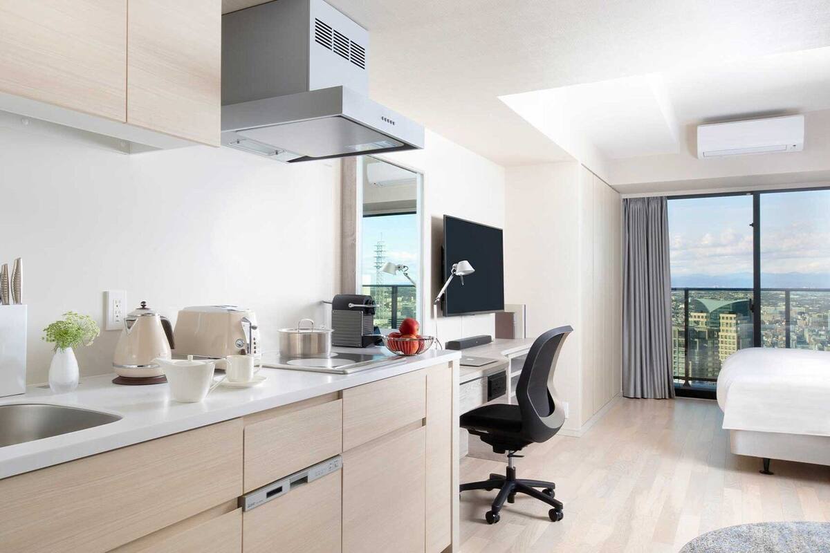 A modern hotel room with a kitchenette, a bed, a desk with a chair, a TV, and a city view.