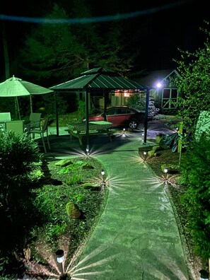 A nighttime outdoor area with a paved walkway, a covered seating area, and a red car parked nearby. - Creekside Cottage - Fallingwater Ohiopyle Seven Springs Laurel Highlands (Normalville)