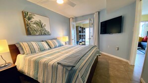 A bedroom with a bed, a nightstand, a ceiling fan, a TV mounted on the wall, and a door leading to another room.