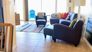 A living room with a sofa, armchairs, a coffee table, a blue chair, a patterned rug, and a large window with curtains.