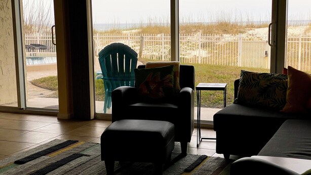 A living room with a view of the beach through large windows, a teal chair, a black sofa, and a small table.