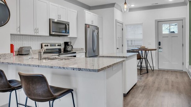 A modern kitchen with white cabinets, a stainless steel refrigerator, a microwave, a coffee maker, and a bar area with stools.