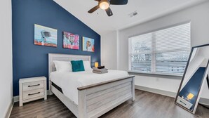 A bedroom with a bed, a nightstand, a ceiling fan, a window with blinds, and artwork on the wall.