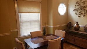 A dining area with a table and four chairs, a window with blinds, a decorative wall piece, and a cabinet with decorative items.