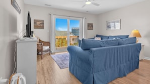 A living room with a sofa, a TV, a ceiling fan, and a view through a sliding door. - Sunset SeaRenity, Turtle Cove 229 | Surf City, NC | Community Pool (Surf City)