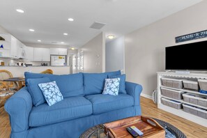 A living room with a blue sofa, a television, and storage shelves. - Sunset SeaRenity, Turtle Cove 229 | Surf City, NC | Community Pool (Surf City)