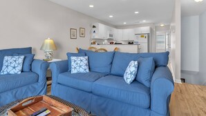 A living room with a blue sofa set, a wooden coffee table, and a kitchen in the background. - Sunset SeaRenity, Turtle Cove 229 | Surf City, NC | Community Pool (Surf City)