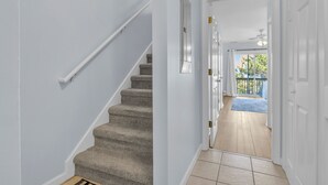 A hallway with a carpeted staircase, a door leading to a balcony, and a view of the outdoors. - Sunset SeaRenity, Turtle Cove 229 | Surf City, NC | Community Pool (Surf City)