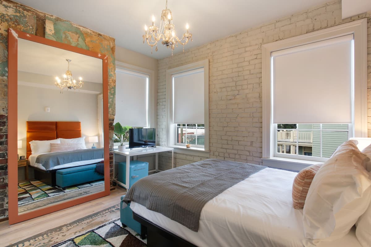A bedroom with a large mirror, a bed with a blue bench, a desk with a TV, and a view of a balcony.