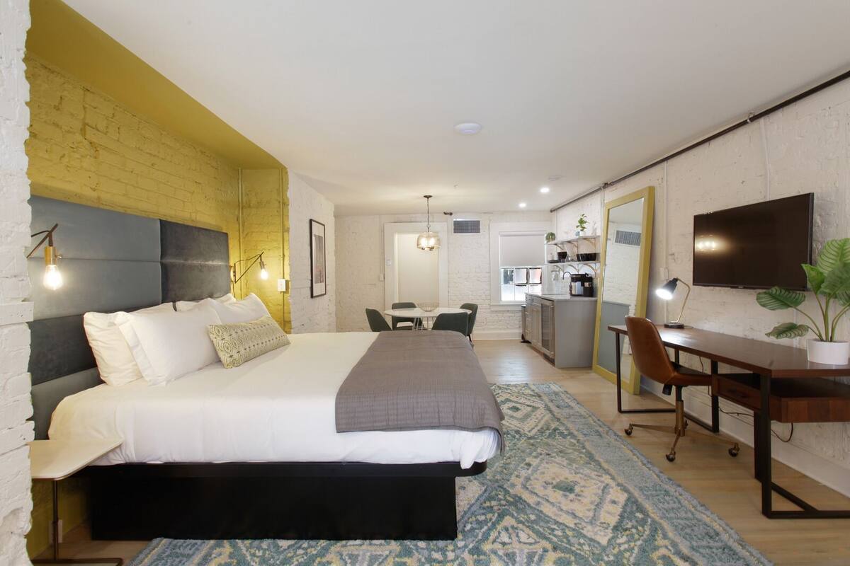 A modern hotel room with a large bed, a desk, and a kitchenette.