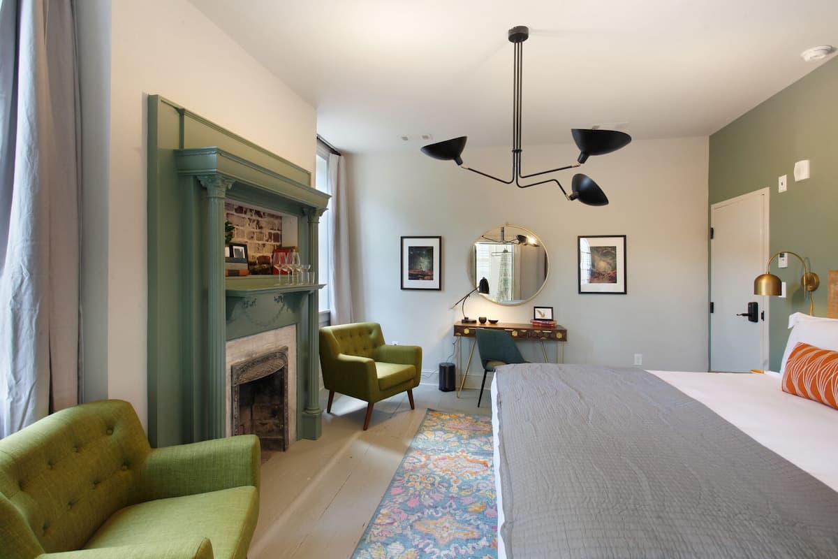 A bedroom with a bed, a desk, two chairs, a fireplace, and a ceiling light fixture.