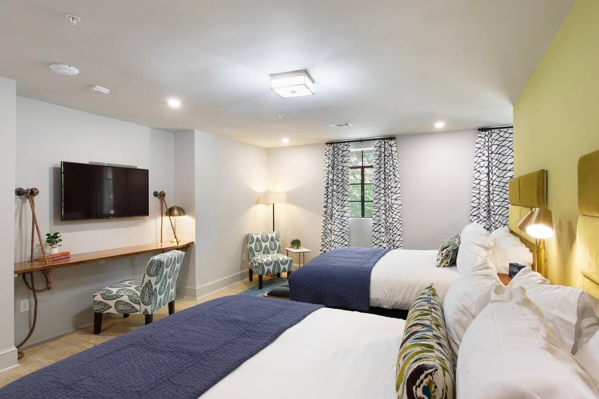 A hotel room with two beds, a TV, a desk, and a window with patterned curtains.