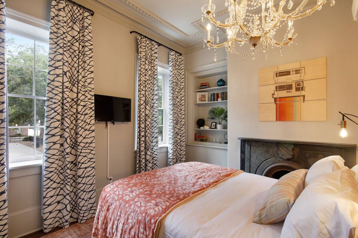 A bedroom with a fireplace, a chandelier, a TV mounted on the wall, and a bookshelf with books and decorative items.