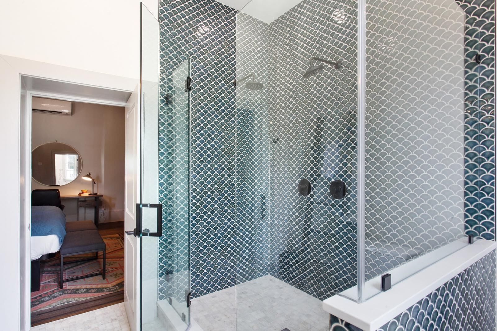 Un baño moderno con cabina de ducha de vidrio, una pequeña zona de estar con una silla y una mesa, y un espejo sobre la mesa.