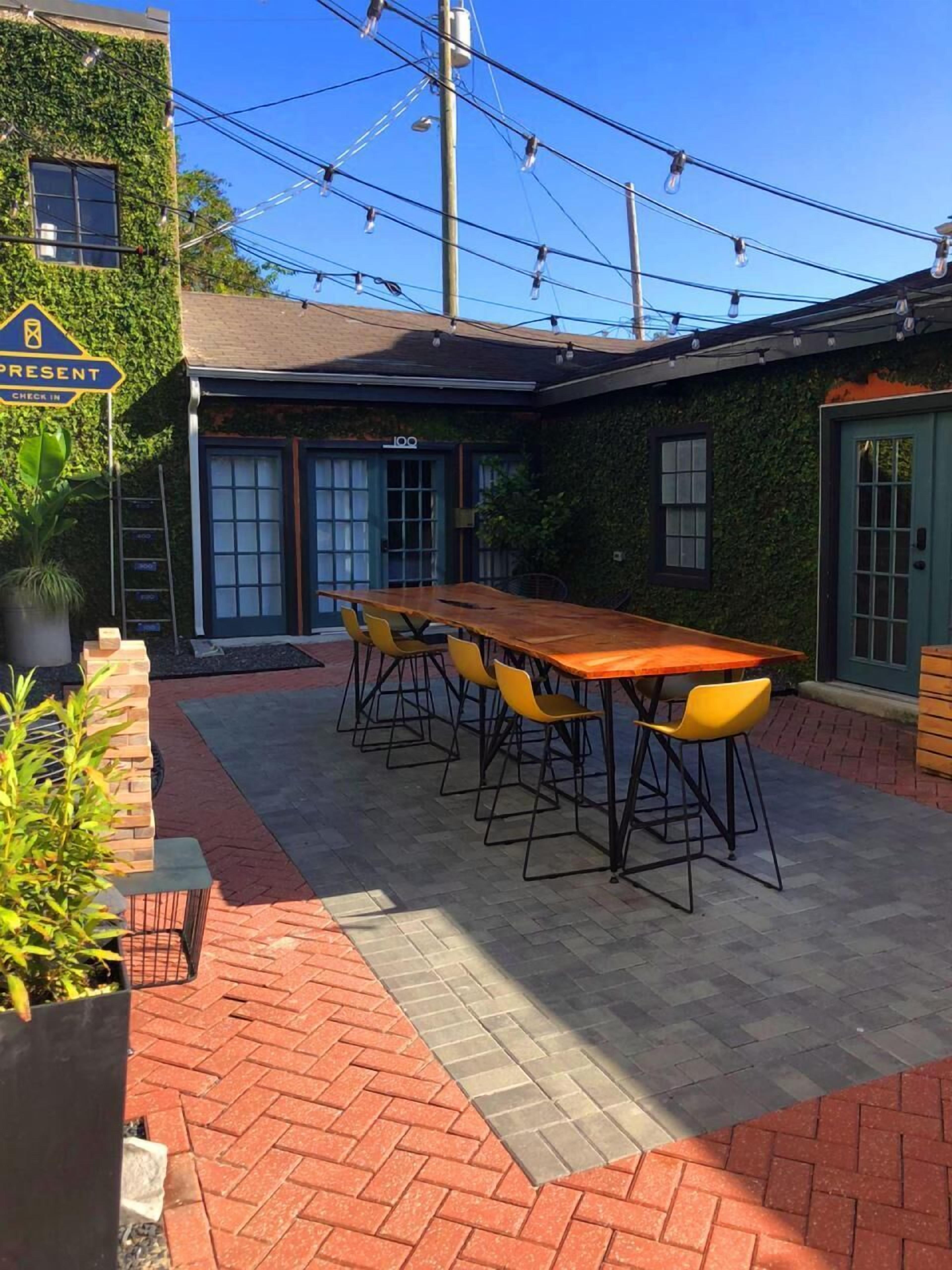 An outdoor patio with a long wooden table and yellow chairs, surrounded by brick flooring and greenery.