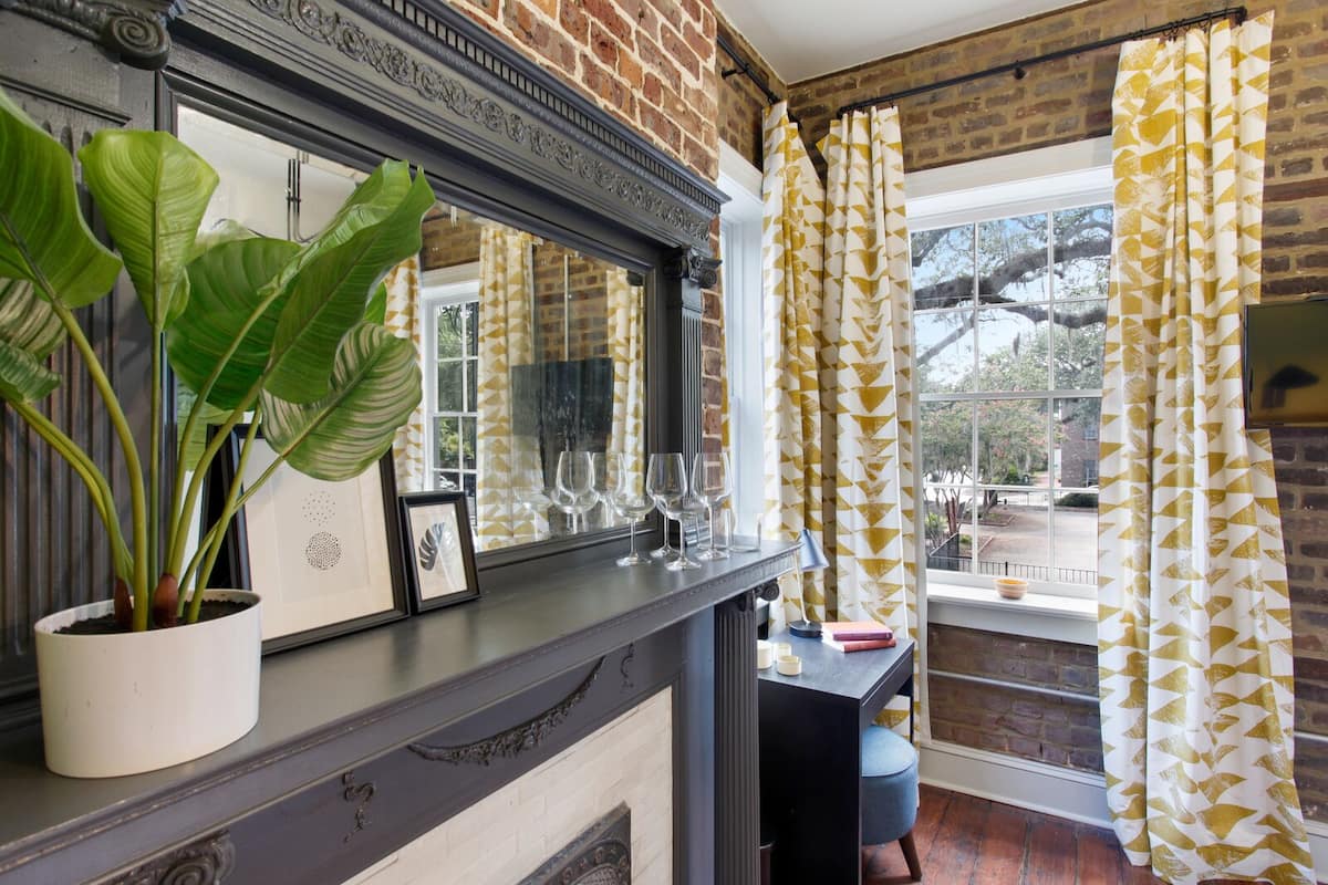 A room with a brick wall, a fireplace, a window with yellow patterned curtains, a plant in a white pot, and a table with wine glasses and a book.
