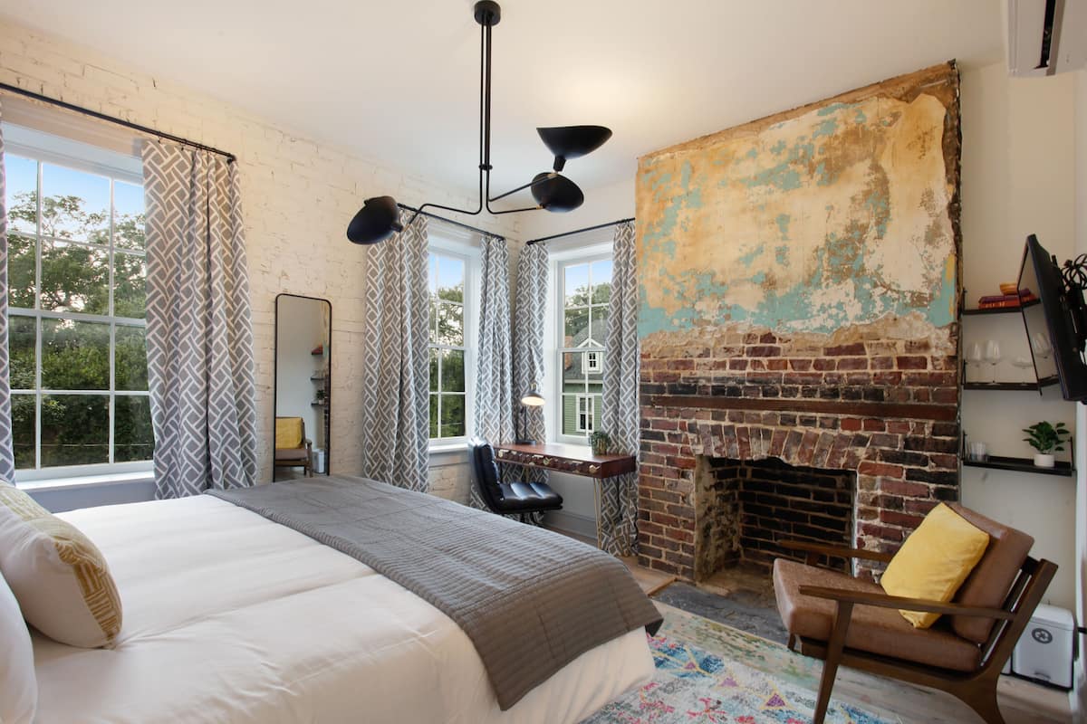 A bedroom with a large bed, a desk, a chair, a fireplace, and a window with curtains.
