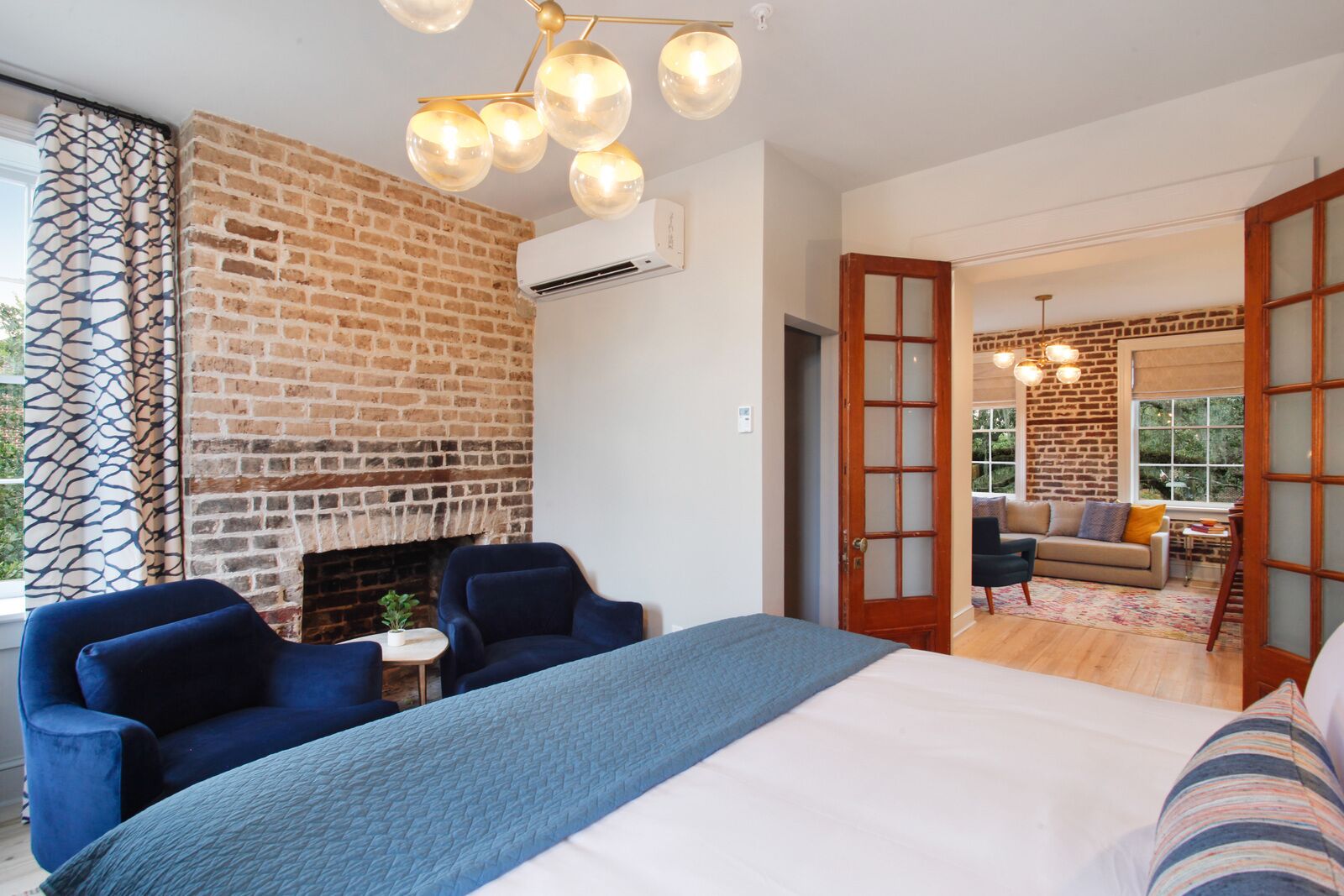 A bedroom with a brick fireplace, a large bed, two blue armchairs, and a small table with a plant.