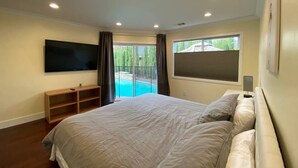 Un dormitori amb un llit gran, un televisor fixat a la paret, una petita estanteria de fusta i una finestra amb vista a una piscina.