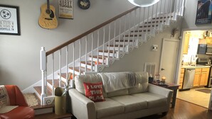 A living room with a beige sofa, a red armchair, a staircase, and a kitchen visible through an open door.