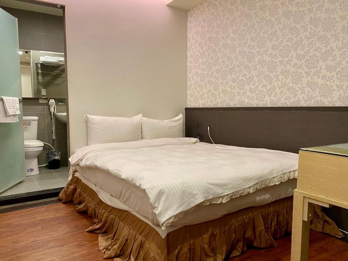 A hotel room with a bed, a wooden floor, a bathroom with a toilet and sink, and a patterned wallpaper on the wall.