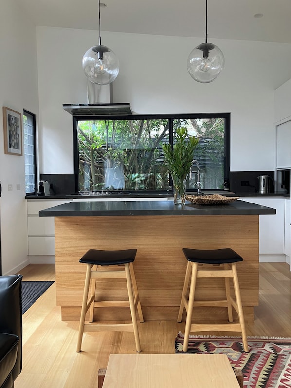 A modern kitchen with a large island, two bar stools, a hanging light fixture, and a view of greenery outside.