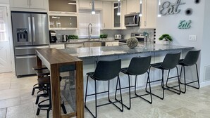 A modern kitchen with a central island, bar stools, stainless steel appliances, and decorative wall art. - Modern Coastal Home near Siesta Key and Downtown (Sarasota)
