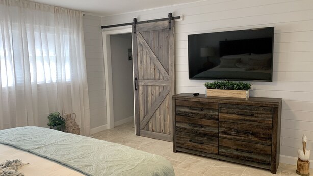 A bedroom with a bed, a sliding barn door, a TV mounted on the wall, and a wooden dresser. - Modern Coastal Home near Siesta Key and Downtown (Sarasota)