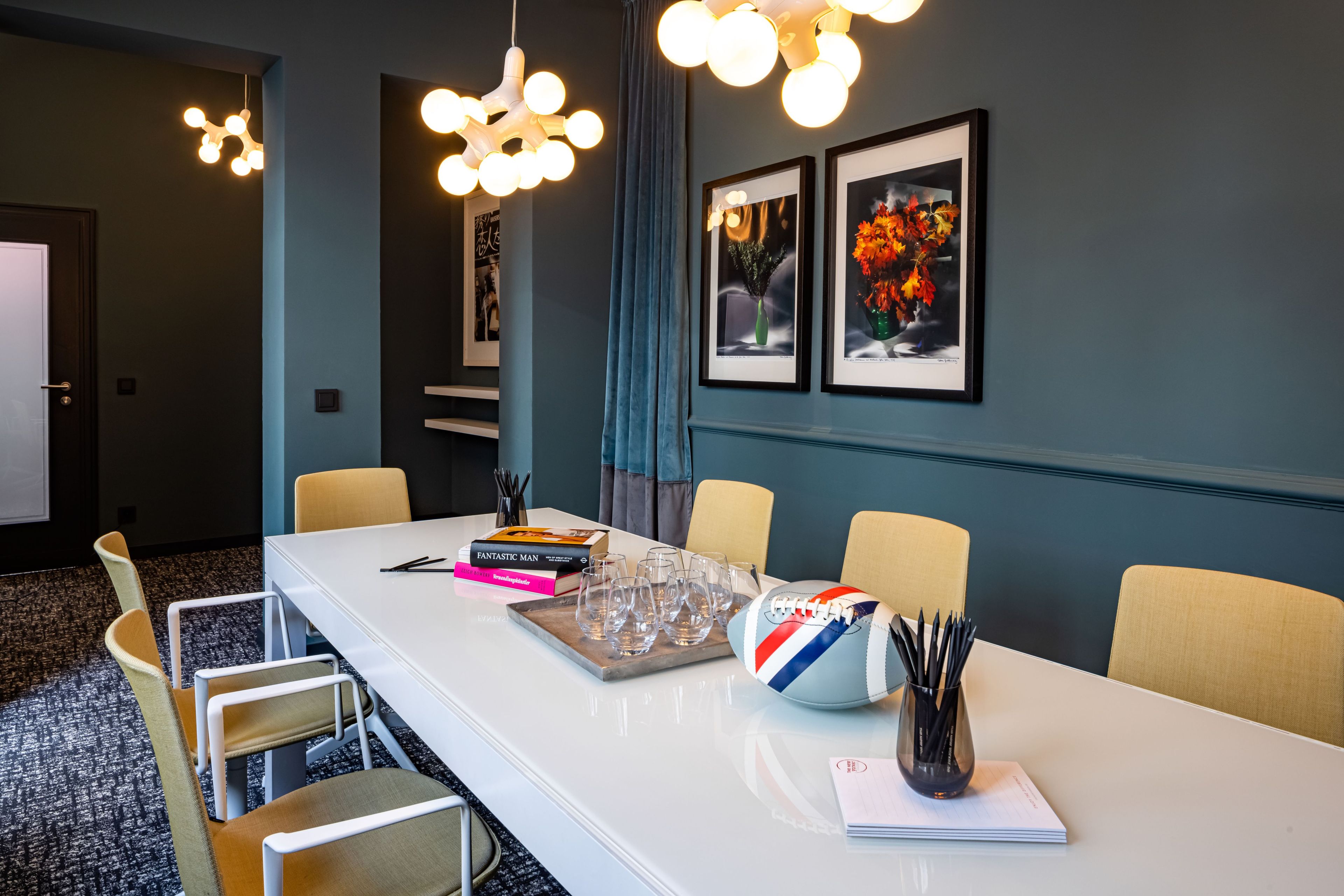 Sala de conferências com mesa branca, cadeiras e uma bola de esportes. Há quadros nas paredes e um candelabro acima.