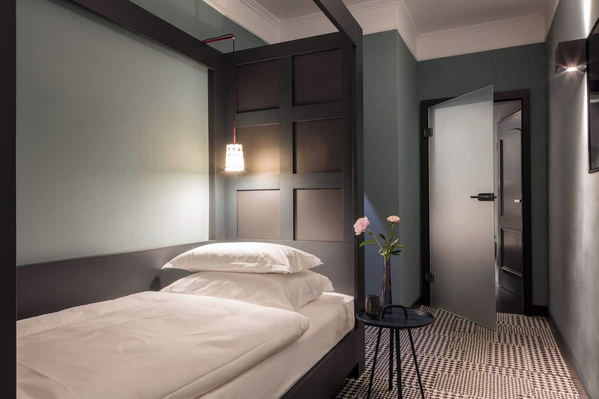A modern hotel room with a neatly made bed, a bedside table with a vase of flowers, and a wall-mounted light fixture.