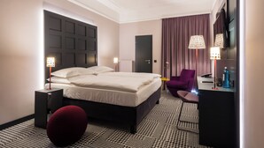 A modern hotel room with a large bed, a small seating area, a flat-screen TV, and a ceiling fan. - The Midtown Hotel (Cologne)