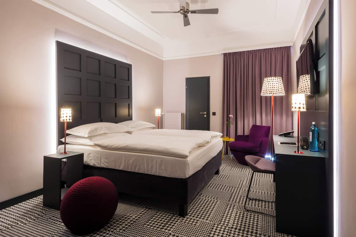 A modern hotel room with a large bed, a ceiling fan, a seating area with a purple chair, and a flat-screen TV.
