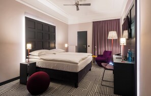 A modern hotel room with a large bed, a small seating area, a flat-screen TV, and a ceiling fan. - The Midtown Hotel (Cologne)