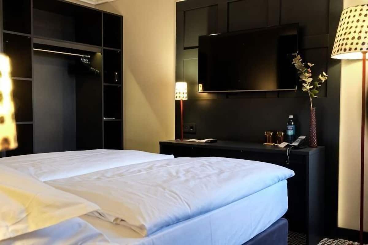 A hotel room with a bed, a TV mounted on the wall, a desk with a lamp, and a wardrobe.