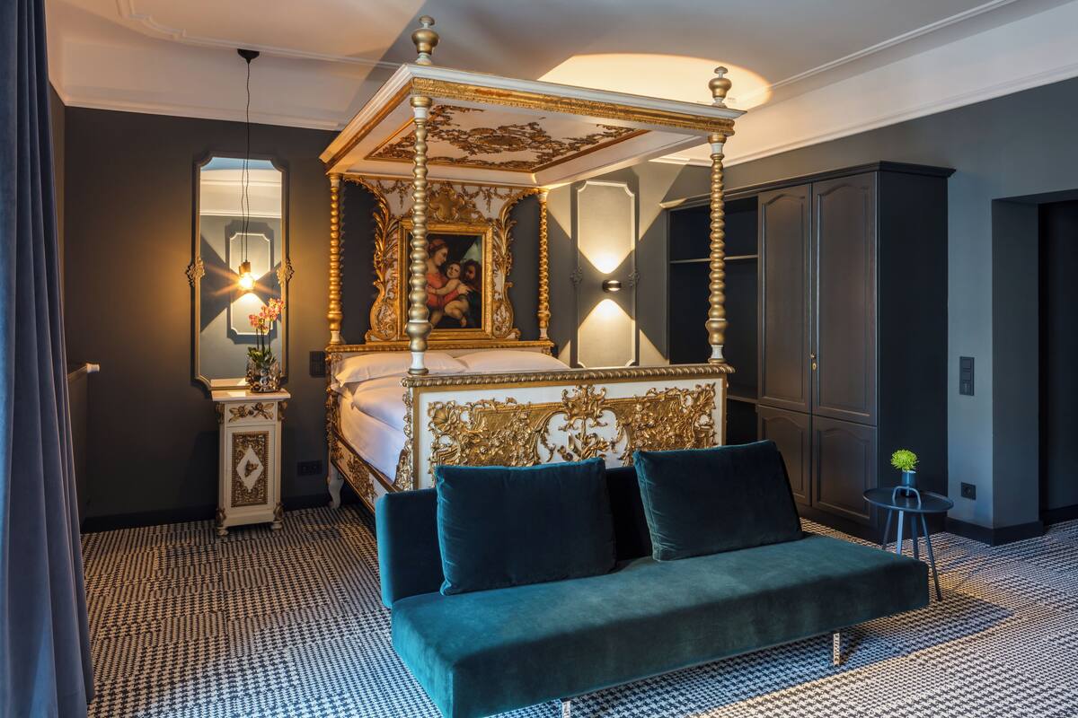 A four-poster bed with a gold and white ornate headboard, a green sofa, a small table with a plant, and a mirror on the wall.