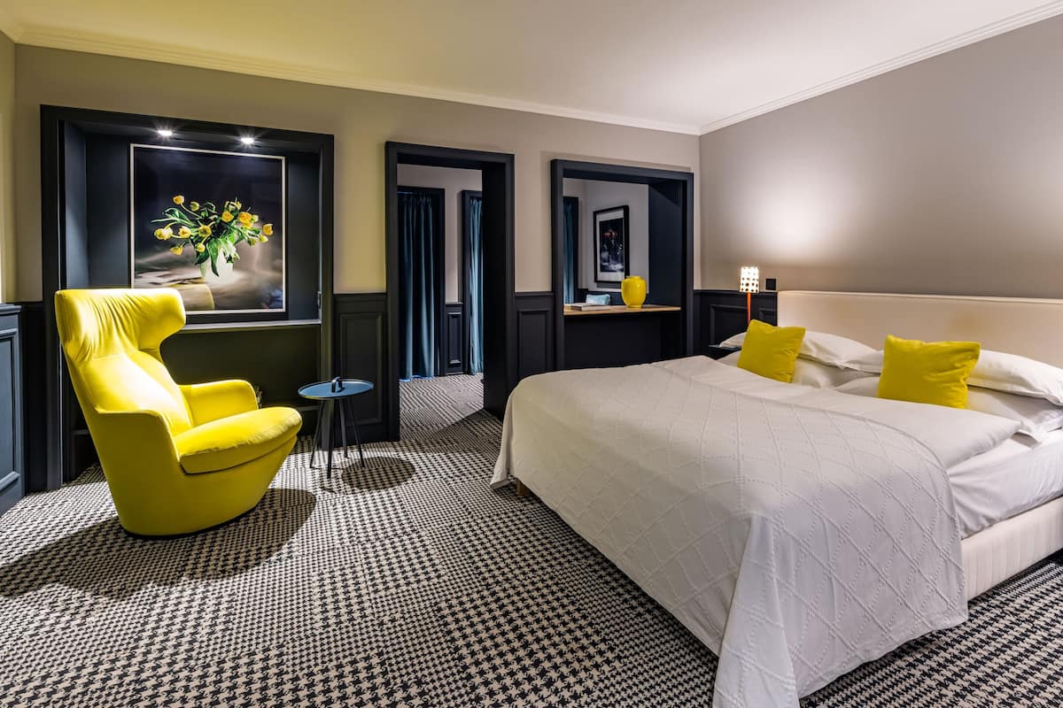 A hotel room with a large bed, a yellow armchair, a small table, and a framed picture on the wall.