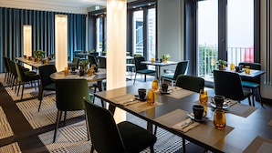 Een modern hotelrestaurant met gedekte tafels voor het ontbijt, grote ramen met uitzicht op de stad, en een balkon met een balustrade.
