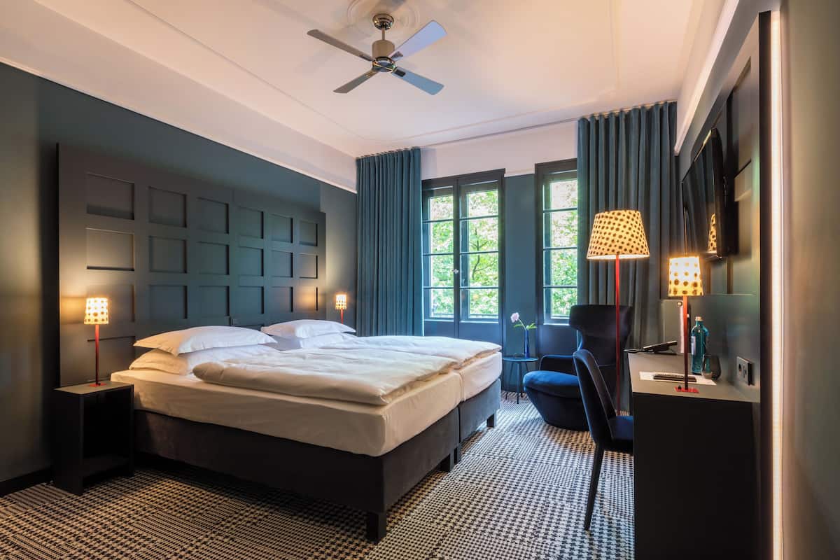A modern hotel room with a large bed, a desk, a chair, and a ceiling fan.