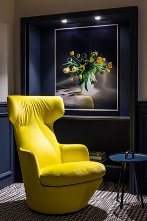 A bright yellow armchair in a room with a framed artwork of yellow flowers on the wall, a small round table, and a patterned carpet. - The Midtown Hotel (Cologne)