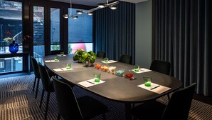 A modern conference room with a long table, black chairs, and a large window with a view of buildings. - The Midtown Hotel (Cologne)