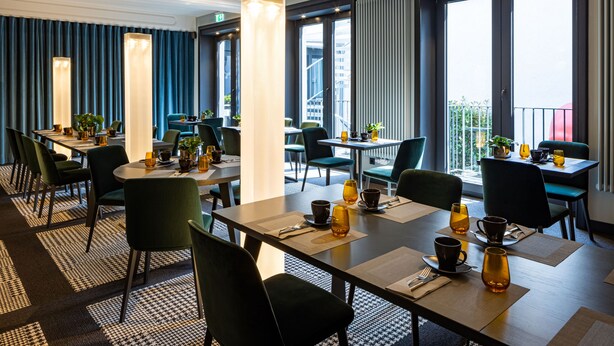 A modern hotel dining area with tables set for breakfast, large windows offering views of the city, and a balcony with a railing. - The Midtown Hotel (Cologne)