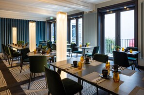 A modern hotel dining area with tables set for breakfast, large windows offering views of the city, and a balcony with a railing. - The Midtown Hotel (Cologne)