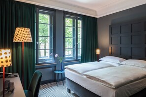 A hotel room with a large bed, two bedside lamps, a desk with a lamp, and a view of the outdoors through two windows. - The Midtown Hotel (Cologne)