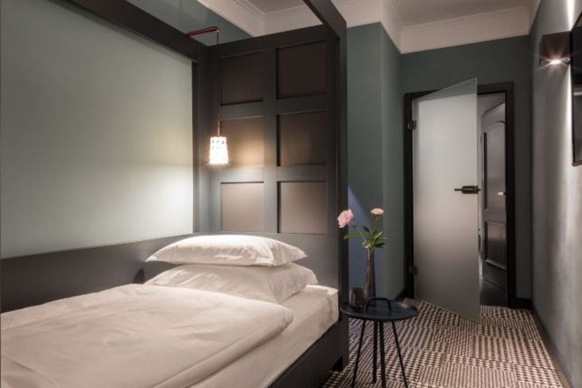 A hotel room with a bed, a bedside table with a vase of flowers, a wall-mounted light, and a door leading to another room.
