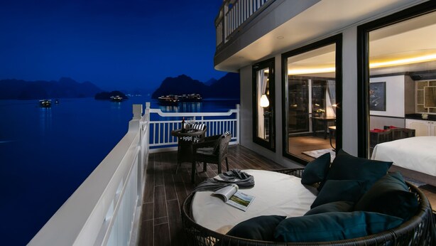 A balcony with a view of the water and mountains, a wicker sofa with pillows, and a table with a lamp. - Lapinta Luxury Cruises (Hai Phong)