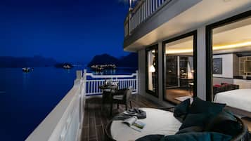 A balcony with a view of the water and mountains, a wicker sofa with pillows, and a table with a lamp.