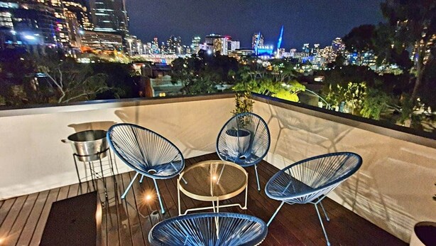 Terraza en la azotea con una mesa y cuatro sillas, con vista a un paisaje urbano nocturno.