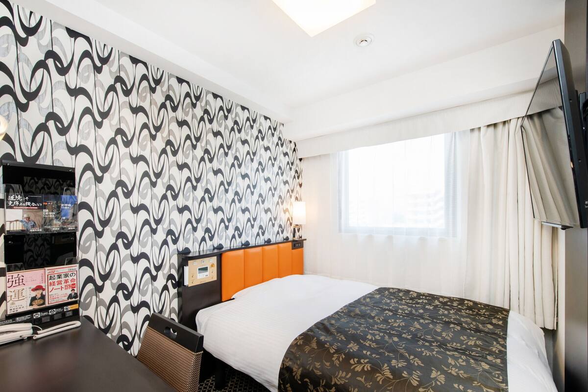 A hotel room with a patterned wallpaper, a bed with a floral bedspread, a desk with a lamp, and a television on the wall.