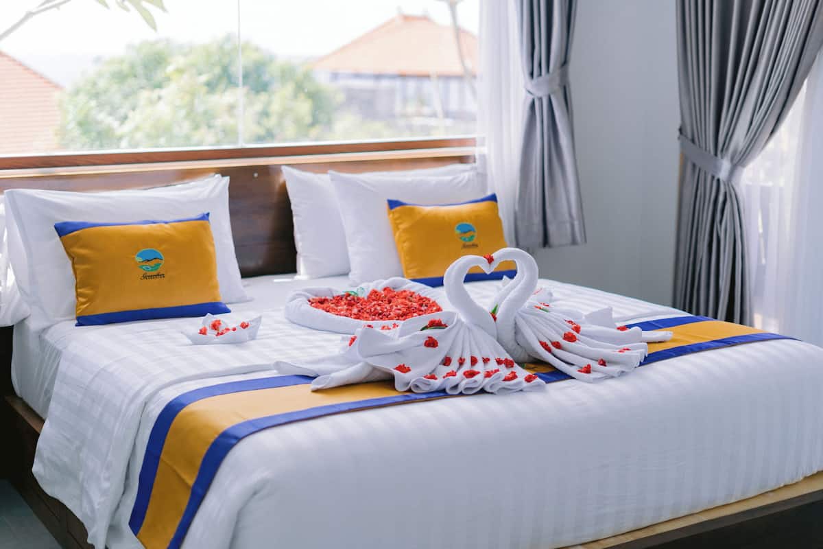 A hotel room with a neatly made bed, two pillows with a logo, a heart-shaped towel arrangement, and a window with curtains.